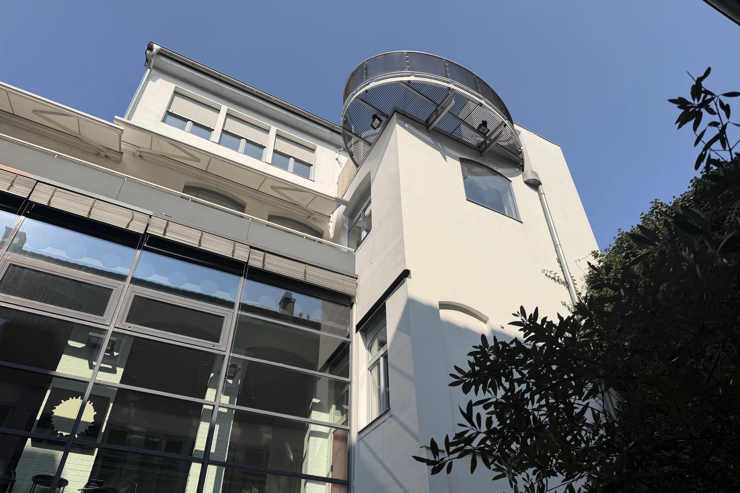 Ovaler Dachbalkon aus Stahl und Glas über dem gläsernen Verbindungsbau, aufgenommen bei blauem Himmel