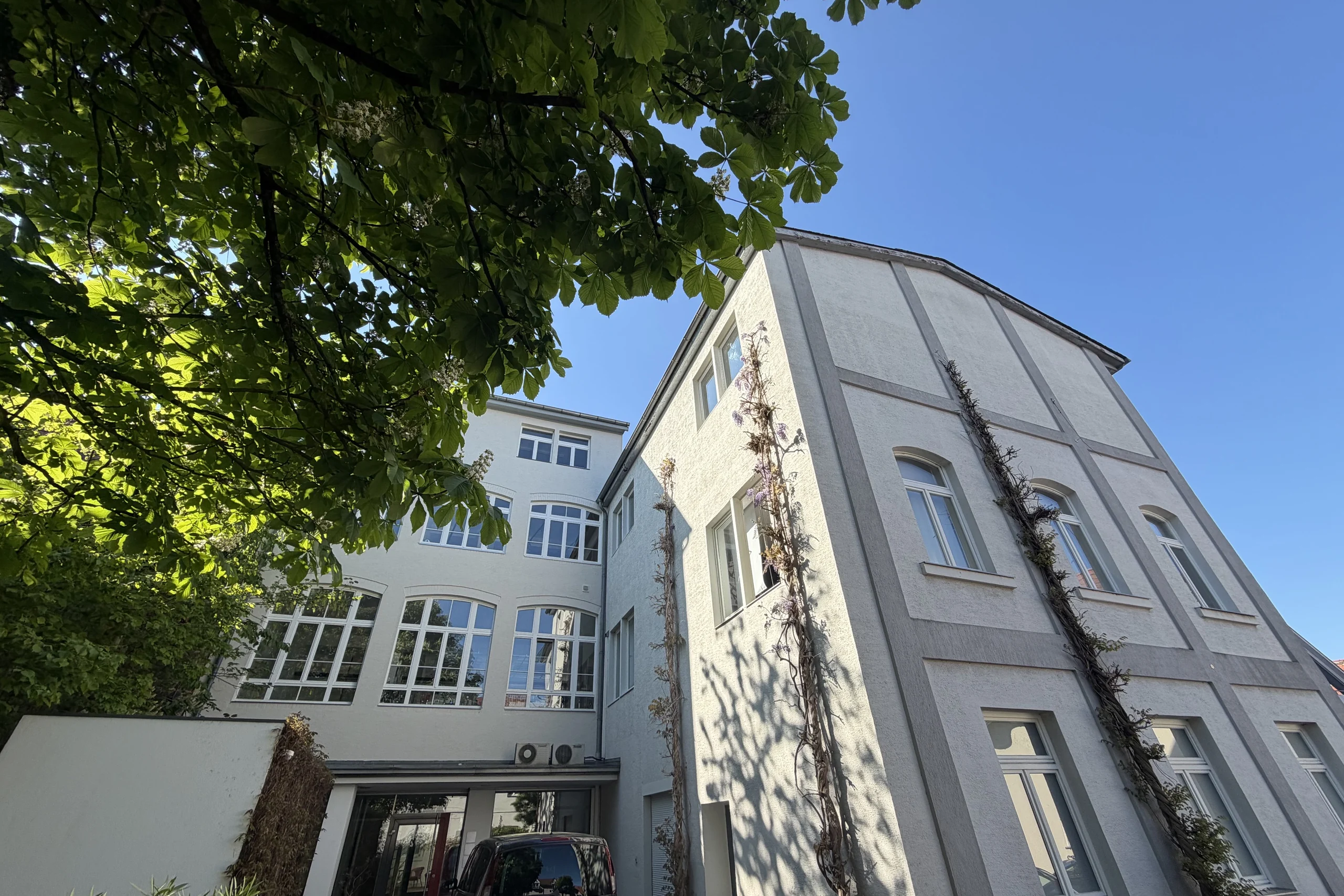 Blick im Hof zwischen den Gebäudeteilen des Ensembles, bei blauem Himmel und belaubten Bäumen